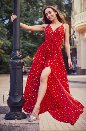 Street fashion: beautiful sweet girl in red dress and sandals stands at lamp post. Portrait of cute smiling young woman on the streetの写真素材