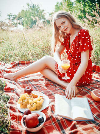 Picnic scene: pretty smiling girl with glass of orange juice sitting on a plaid and reading book in park. Outdoor portrait of beautiful young woman in red dress. Effect of old film with grainの写真素材