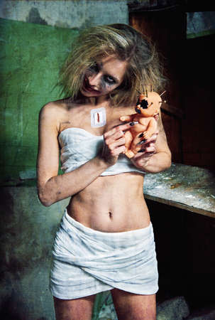 Strange mad girl with creepy ugly doll in forsaken place (asylum). Crazy young woman (freak) dressed in messy bandages. Portrait of the smiling madman (mental sick). Grunge texture effectの写真素材