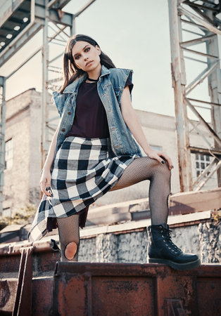 Outdoor portrait of pretty grunge (rock) girl at industrial place. Informal model dressed in jean jacket, checkered shirt, boots and holey tightsの写真素材
