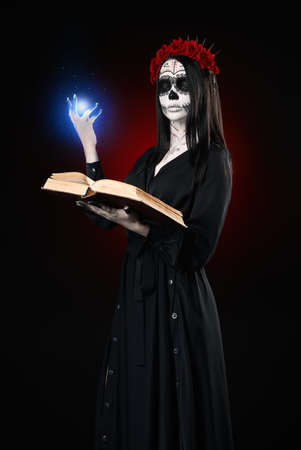 Young woman with calavera (sugar skull) makeup holds book (grimoire) and casts the spell. The witch girl with muertos (muerte) greasepaint in black dress. Day of the dead (and Halloween) themeの写真素材