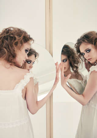 Portrait of lovely strange freaky girl. Beautiful curious woman in white dress with mirror in hands standing at mirrorの写真素材