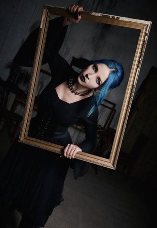 Indoors portrait of lovely goth girl in black dress. Blue-haired gothic lady holds a picture frame...
