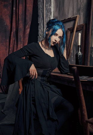 Indoors portrait of lovely goth girl in black dress. Blue-haired gothic lady sitting on chair at the table. vintage lookの写真素材