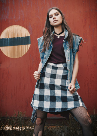 Outdoor portrait of the pretty grunge (rock) girl standing at the wall. Informal model dressed in jean jacket, checkered shirt and holey tightsの写真素材