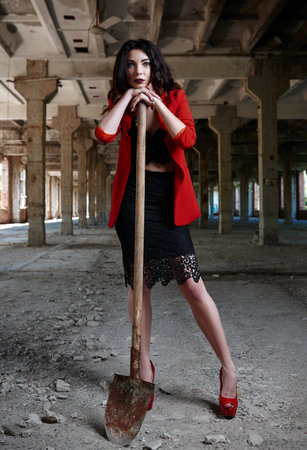 Portrait of beautiful girl with shovel in hands. Cute young woman dressed in red jacket, black skirt and brassiere among forsaken industrial placeの写真素材