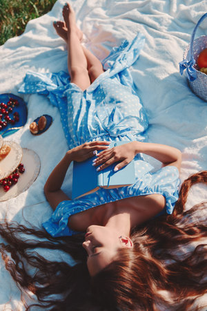 Picnic scene: pretty girl sleeping on a plaid with book in hands. Outdoor portrait of cute young woman in blue dressの写真素材