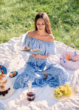 Picnic scene: pretty girl sitting on a plaid and reading book in park. Outdoor portrait of beautiful young woman in blue dressの写真素材
