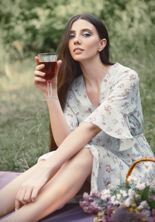 Picnic scene: cute sensual girl sitting on a plaid in park and drinking juice (wine). Outdoor portrait of beautiful young woman in dress. Effect of old faded film. vintage lookの写真素材