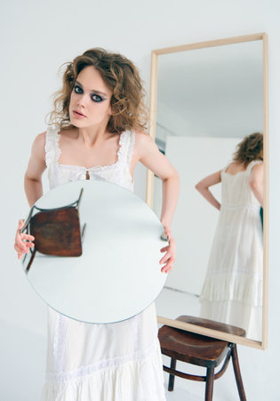 Portrait of lovely strange freaky girl. Beautiful odd woman in white dress with a mirror in handsの写真素材