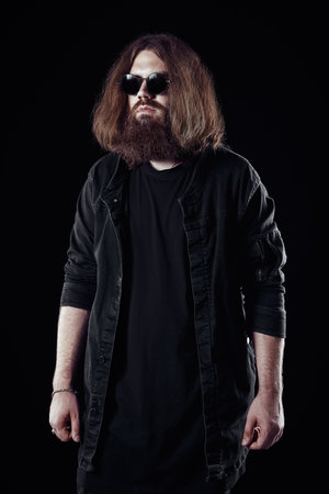 Studio fashion: handsome young man wearing jeans, coat and sunglasses. Portrait of informal long-haired and bearded guy against black backgroundの写真素材