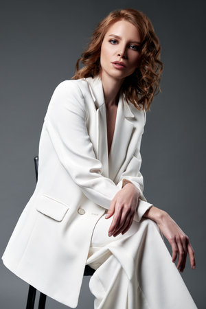 Studio fashion shot of the adorable young woman. Portrait of pretty girl dressed in white suit (jacket and pants). Beautiful lady sitting on chair against gray backgroundの写真素材