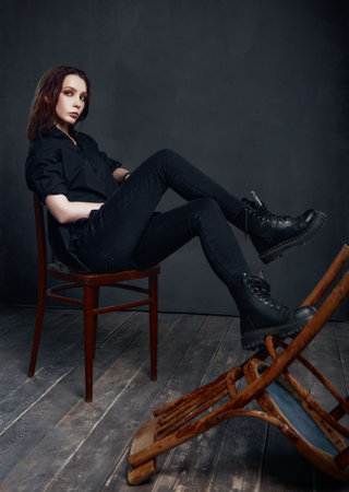 Indoors portrait of informal (rock) girl sitting on a stool. Beautiful young woman in black shirt, jeans and boots. Full lengthの写真素材
