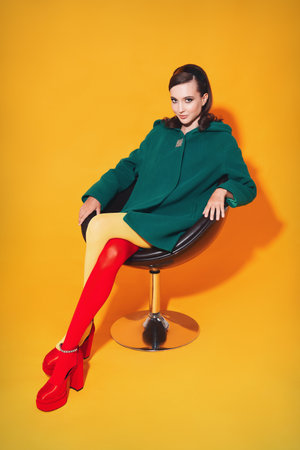Sweet smiling girl in sixties style. Retro (vintage) portrait of charming stylish young woman in coat, shoes and colorful stockings sitting in chair, against yellow background. 60's look dressed ladyの写真素材