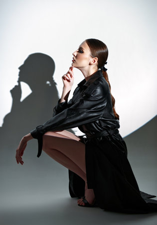 Studio portrait of beautiful young woman sitting on a floor. Elegant lovely girl dressed in black leather trench coat. Round gobo light effect. Profile viewの写真素材