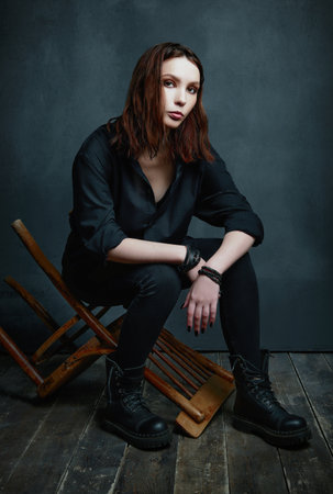 Indoors portrait of informal (rock) girl sitting on a chair. Beautiful young woman in black shirt, jeans and boots. Full lengthの写真素材
