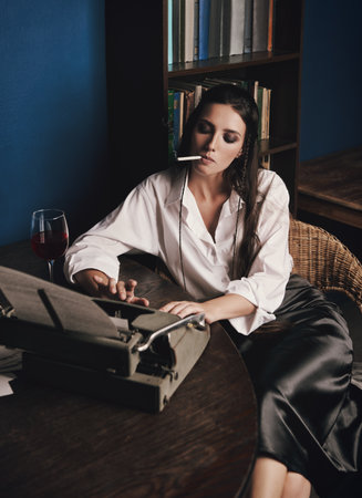 Writer (journalist) typing on old typewriter, smoking cigarette and drinking wine. Indoors old-fashioned portrait of attractive young girl. Retro (vintage) styleの写真素材