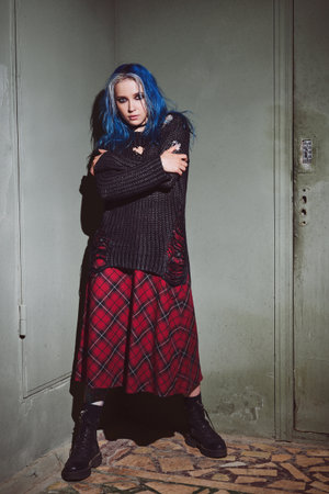 Indoor portrait of cool grunge (rock) girl. Informal blue-haired model, dressed in checkered skirt, and holey sweater standing at the wall. Full lengthの写真素材