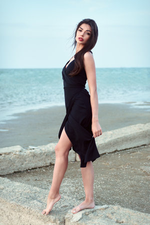 Portrait of lovely young woman in black dress on the ocean coast. Elegant beautiful girl on the beach at sunset timeの写真素材