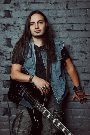 Young rock musician (metalhead) with an electric guitar. Portrait of a handsome long-haired man standing at wall. Indoor shotの写真素材