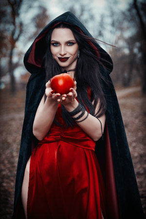 The insidious and seductive young witch offers a treat in the form of a poisoned red apple. Portrait of the cunning smiling sorceress in an autumnal forestの写真素材