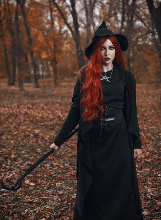 Halloween (Samhain) theme: a ginger (red-haired) witch with horned staff in the forest. Portrait of beautiful redhead enchantress in autumnal woodsの写真素材