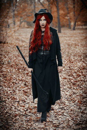 Halloween (Samhain) theme: a ginger (red-haired) witch with horned staff going by the forest. Portrait of beautiful redhead sorceress in autumnal woodsの写真素材