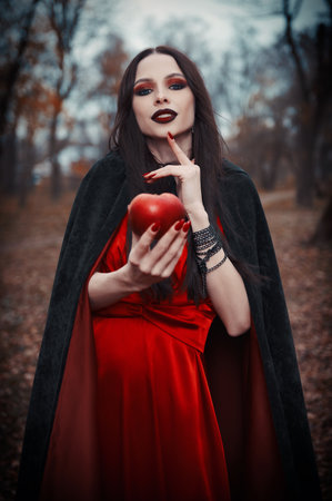 The insidious and seductive young witch offers a treat in the form of a poisoned red apple. Portrait of the smiling sly sorceress in an autumnal groveの写真素材