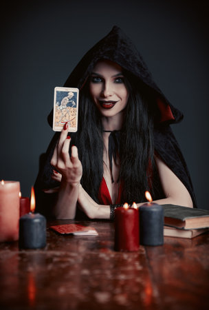 Dark beauty: lovely young fortune teller showing tarot card. Portrait of smiling witch. Beautiful diviner in black robeの写真素材