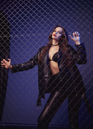 Informal (rock) girl dressed in black leather jacket, pants and bra. Portrait of the beautiful young woman standing behind metallic gridの写真素材