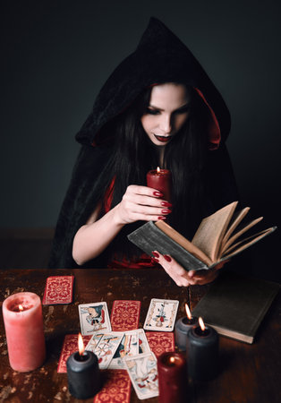 Dark beauty: mysterious young sorceress reading the spellbook. Portrait of witch with book and candle. Lovely soothsayer in black robeの写真素材