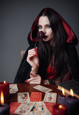 Dark beauty: gorgeous young sorceress sniffs a withered rose. Portrait of smiling witch. Pretty diviner in black robeの写真素材