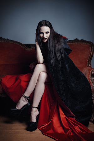 Dark beauty: dangerous young woman in image of witch sitting on sofa. Portrait of smiling girlの写真素材