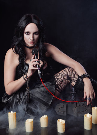 Attractive informal (metalhead) girl singing in microphone. Portrait of female rock (goth) singer sitting on a stage among candlesの写真素材