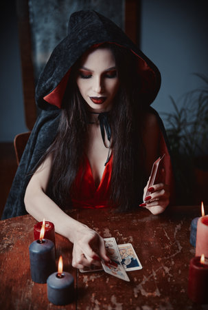 Dark beauty: lovely young soothsayer reading tarot cards. Gorgeous sorceress making Tarot layout. Beautiful witch in black cloakの写真素材