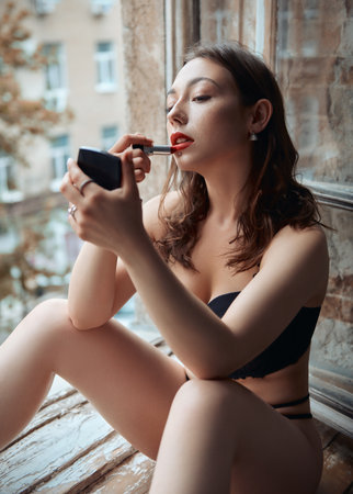 Beautiful young woman sitting on the windowsill with lipstick and mirror and painting her lips. Portrait of attractive girl in black underwearの写真素材
