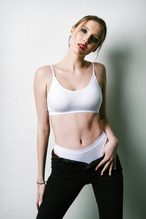 Studio fashion: pretty girl in black jeans and white lingerie. Seductive young woman stands at the wallの写真素材