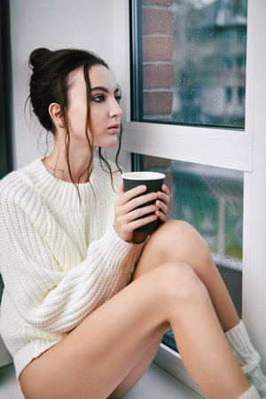 Indoor portrait of beautiful girl dressed in sweater. Sad young woman sitting with cup of coffee by the window on a rainy dayの写真素材