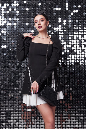 Indoors fashion: portrait of gorgeous young woman in black and white dress with handbag. Charming girl against shiny metallic backgroundの写真素材