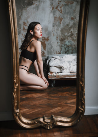 Portrait of lovely girl in black lingerie. Beautiful young woman sitting on floor at the mirror in old roomの写真素材