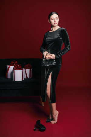 New Year and Christmas fashion: pretty young woman in black dress with glass of champagne (sparkling wine) and purse in hand. Studio portrait of adorable girl standing near giftboxesの写真素材