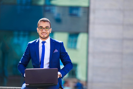 Handsome young Middle Eastern businessman in classic suit holding a laptop and smiling near office buildingの写真素材
