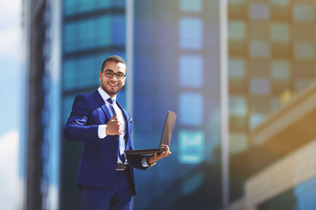 Competent business expert. Confident young handsome man in suit holding laptop and smiling while standing against office buildingの写真素材