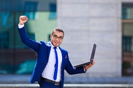 Excited young businessman with laptop keeping arm raised and expressing positivity while standing outdoors with office building in the background. Celebrating successの写真素材