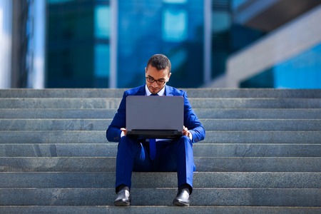 Handsome businessman in suit and eyeglasses working on laptop sitting on stairs near office buildingの写真素材