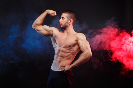Strong young man bodybuilder demonstrating huge muscles on smokeの写真素材