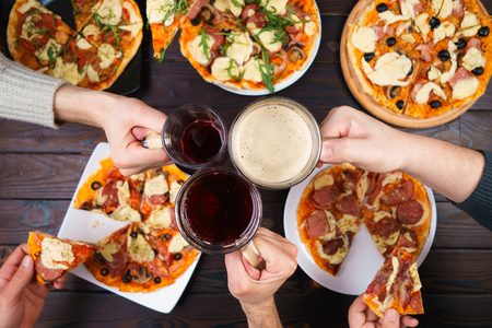 Friends eating pizza. Top view on male hands clinking mugs with beer over table with many pizzas. Party, friendship, pub, restaurant conceptの写真素材