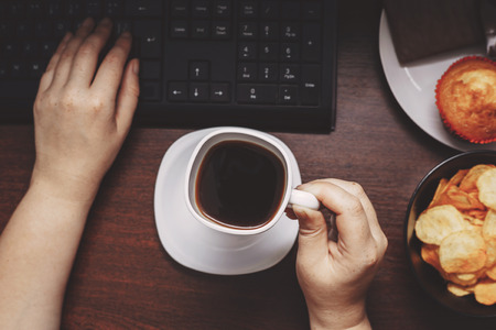 Woman with coffee eating junk food at workplaceの写真素材