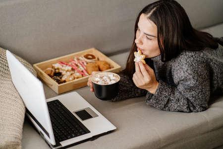Woman eating sweets watching tv series on laptopの写真素材