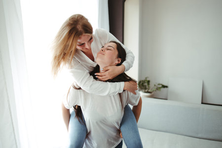 Happy playful girlfriends in love sharing time together indoors giving a piggyback hug. Human...